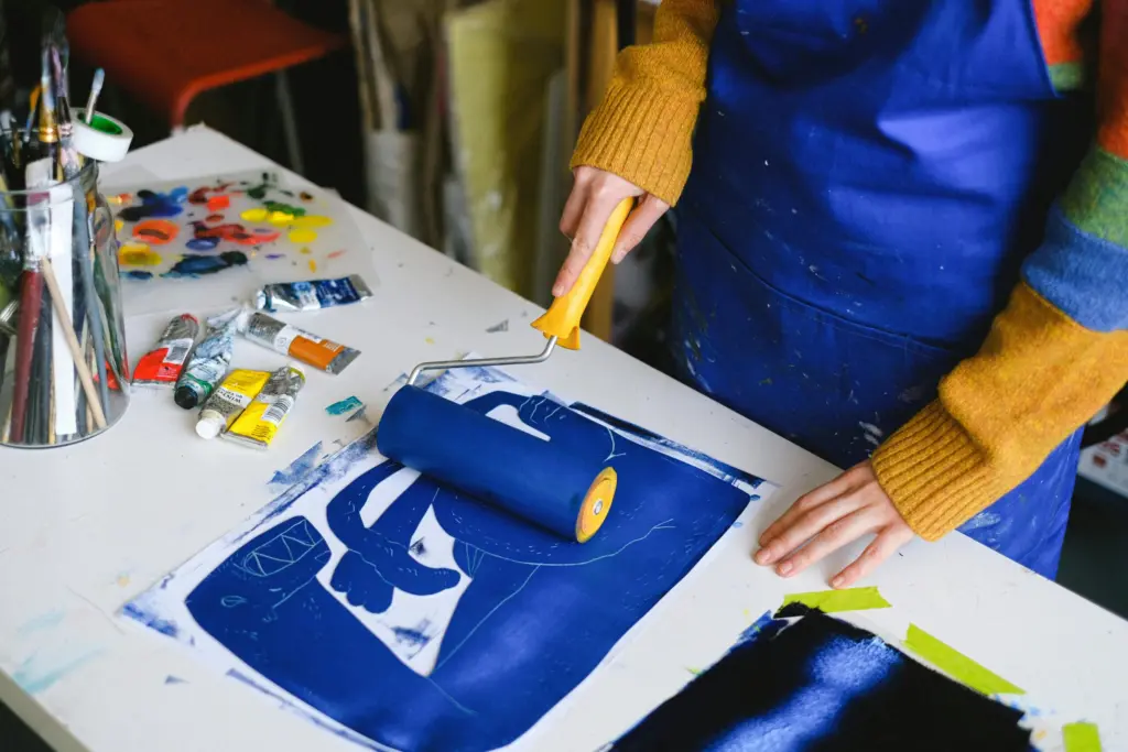 High angle of unrecognizable female artist in apron using paint roller for colouring picture on paper in craft shop